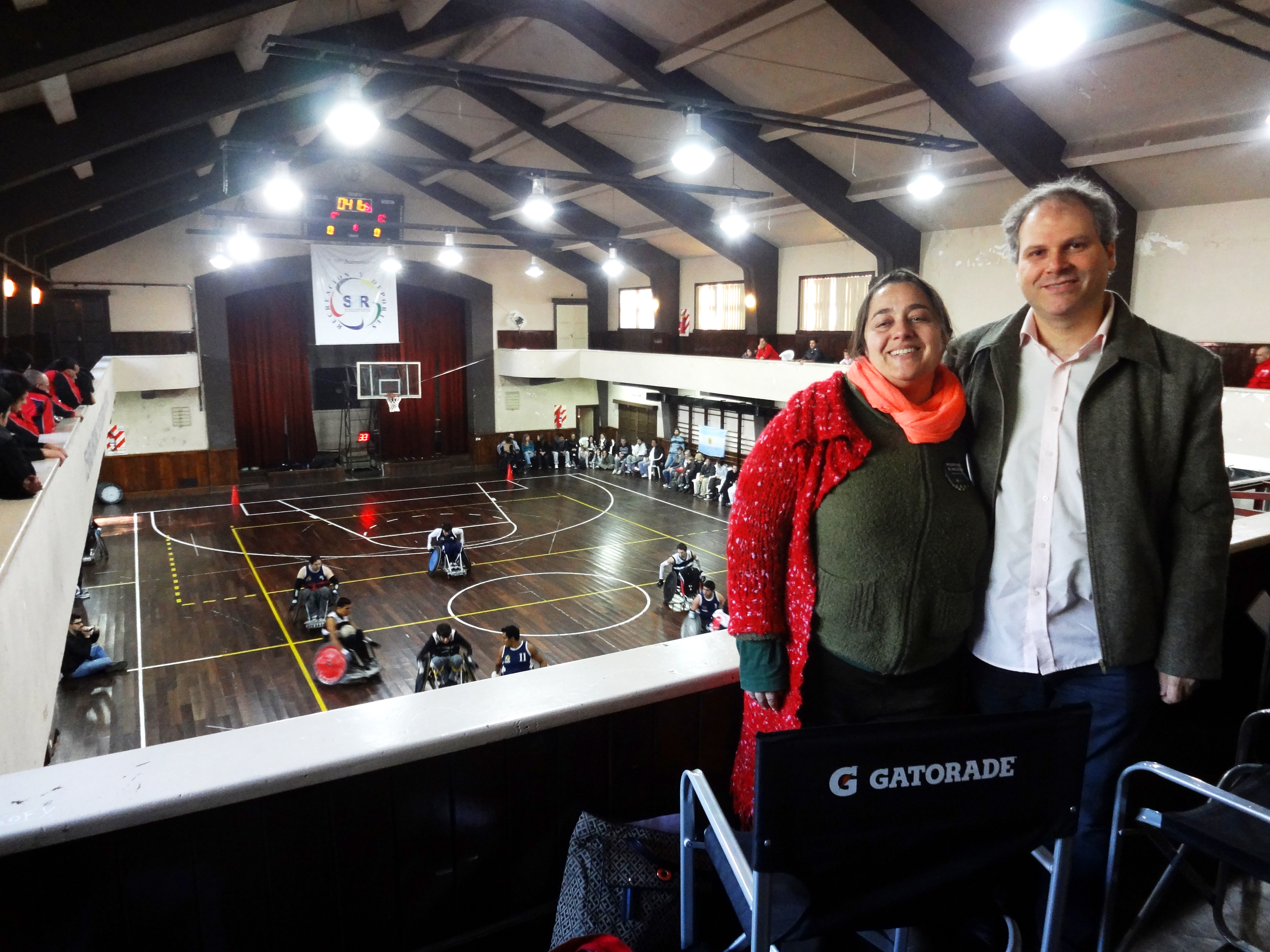 Homem e mulher posam para foto, em pé, no canto esquerdo da imagem. Eles estão na parte superior de um ginásio. Na quadra, de piso de madeira, atletas jogam basquete em suas cadeiras de rodas.