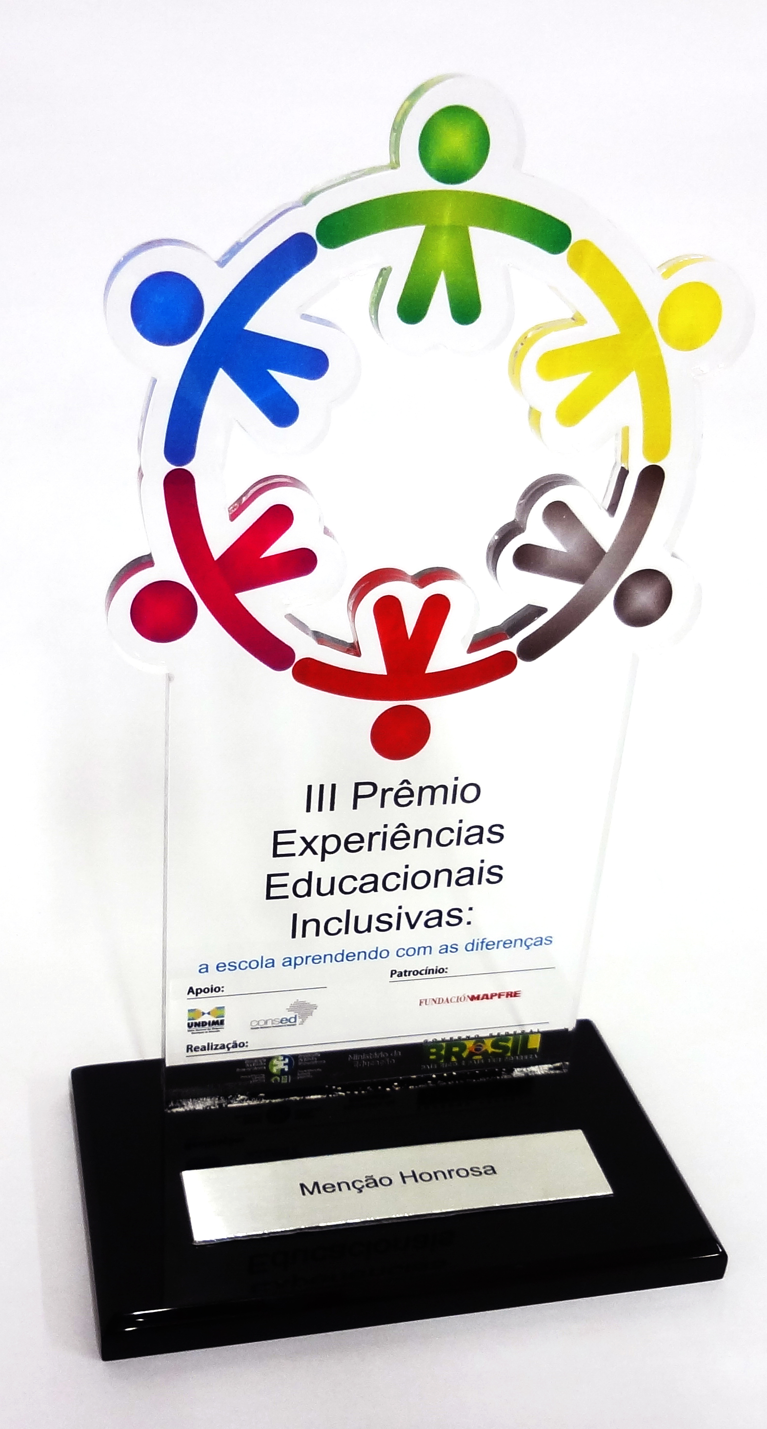 Troféu do prêmio em acrílico, com símbolos coloridos de pessoas de mãos dadas formando um círculo. Em sua base, uma placa com a descrição “Menção Honrosa”.