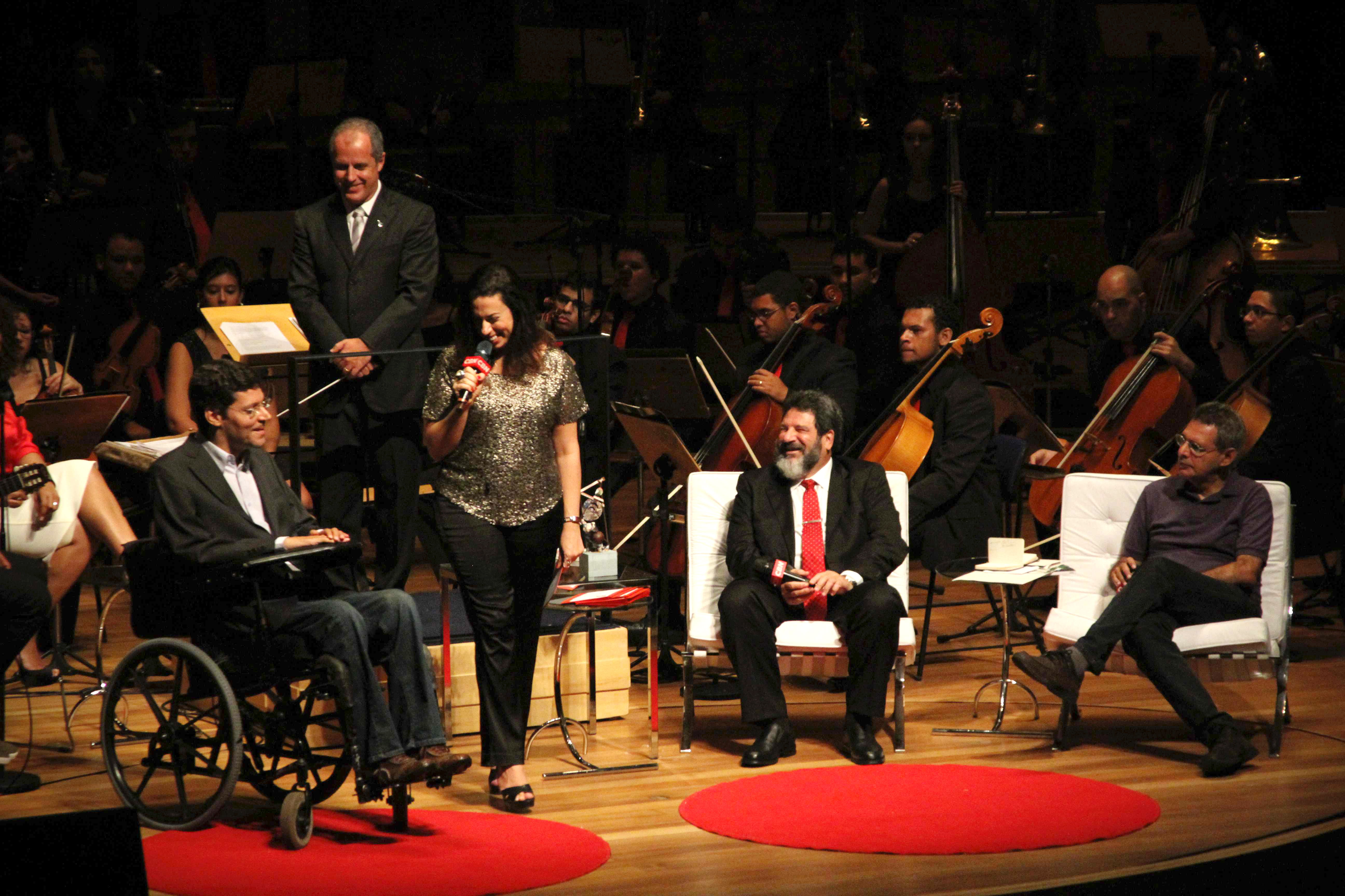 Em um palco, Rodrigo em sua cadeira de rodas, está ao lado de mulher que segura microfone. Ao lado direito da imagem, dois homens sentados em poltronas. Ao fundo maestro e orquestra. Todos sorriem e olham para Rodrigo.
