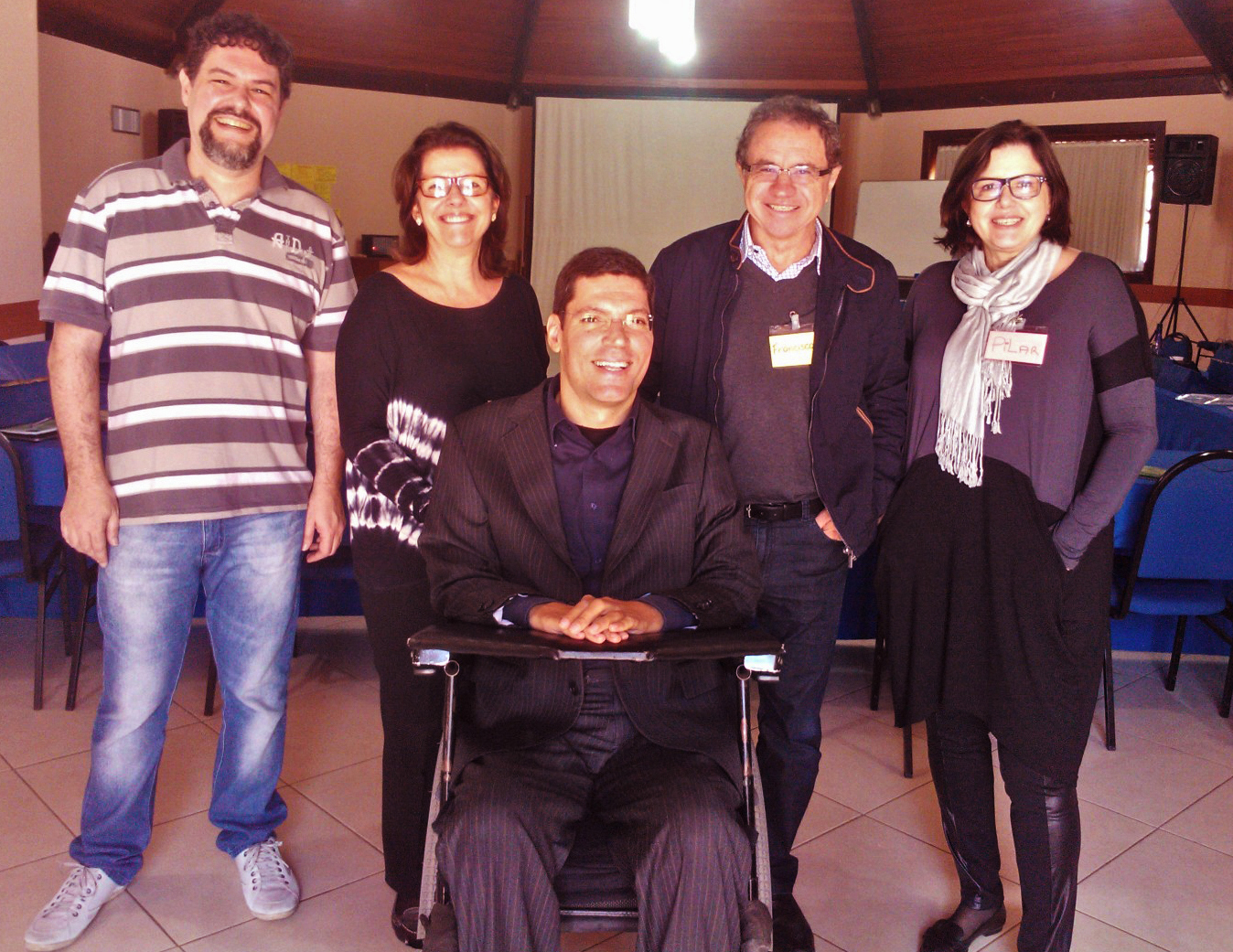 Cinco pessoas posam sorridentes para fotografia em auditório. Eles estão em pé e Rodrigo ao centro em cadeira de rodas.