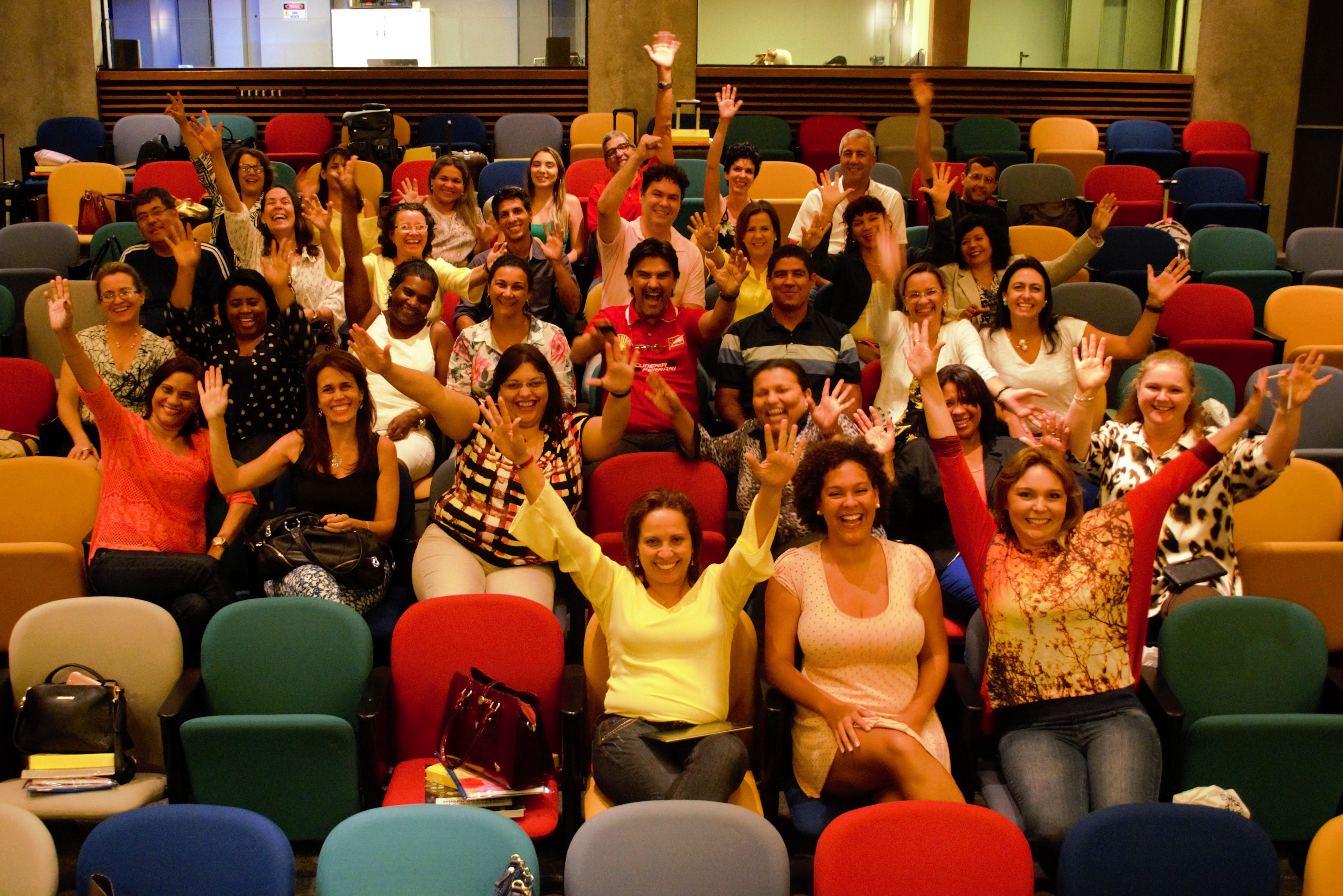 : Homens e mulheres animados e com os braços erguidos posam para foto em auditório com cadeiras coloridas.