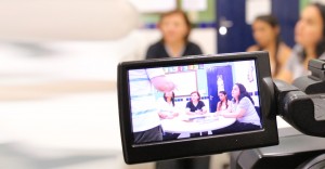 Por um visor de uma câmera filmadora, está um grupo de pessoas conversando ao redor de uma meda redonda em uma sala de aula