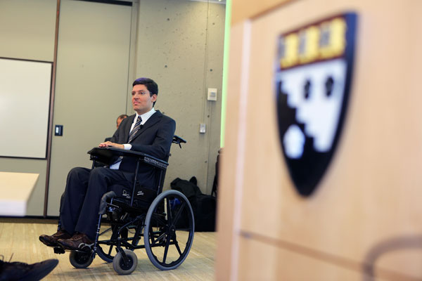 Porta entreaberta com brasão de Harvard. Dentro da sala, Rodrigo está em sua cadeira de rodas.