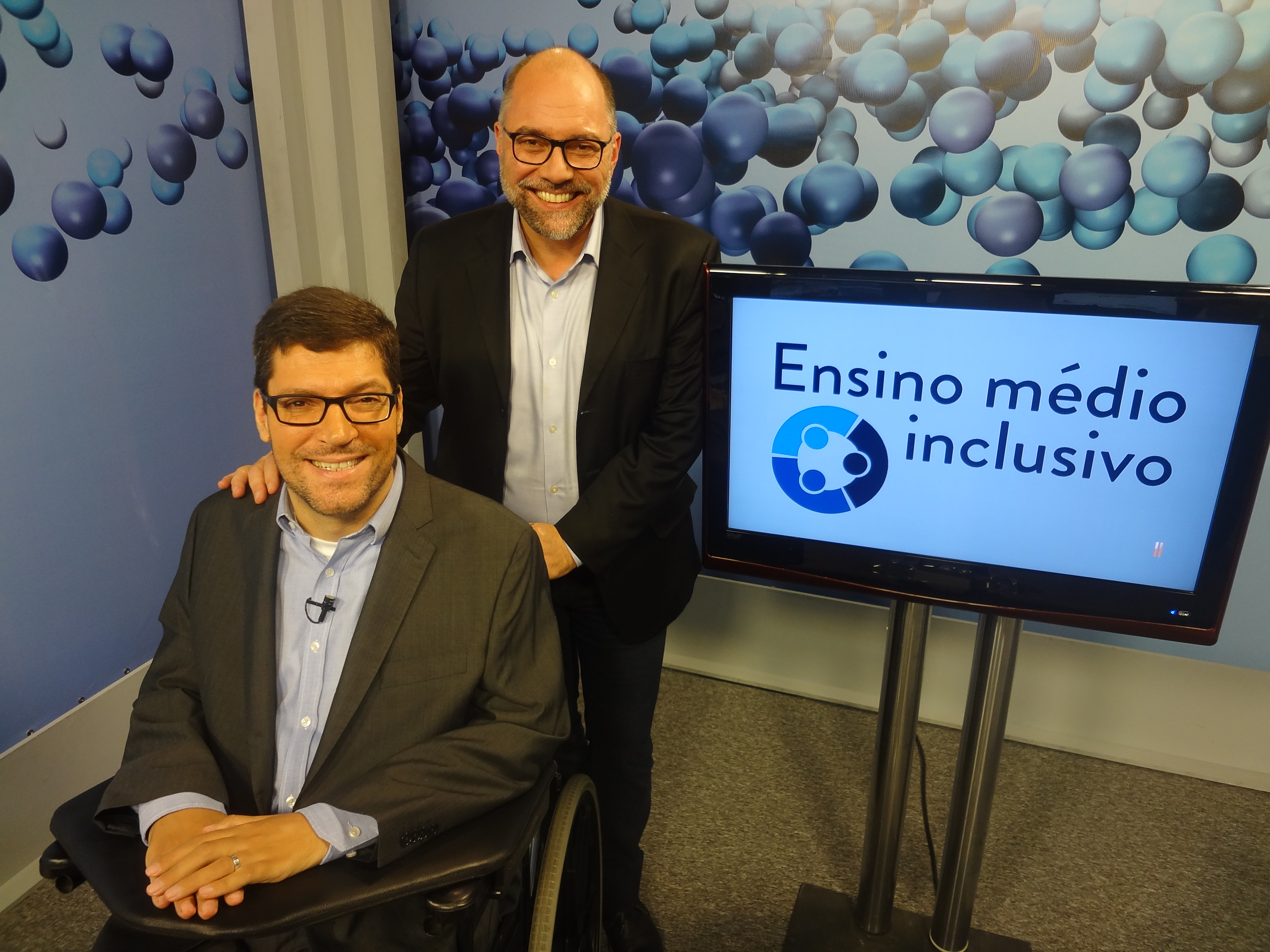 Rodrigo Hübner Mendes e Ricardo Henriques sorriem, ao lado televisão com o logo do Ensino médio inclusivo