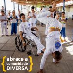 Homem joga capoeira com jovem em cadeira de rodas. Ao redor, crianças e adultos batem palma. Texto: Abrace a Comunidade DIVERSA.