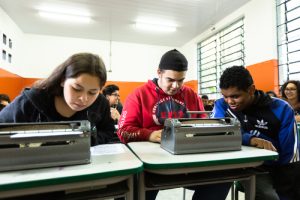 Três adolescentes usam duas máquinas de braille dentro de uma sala de aula.
