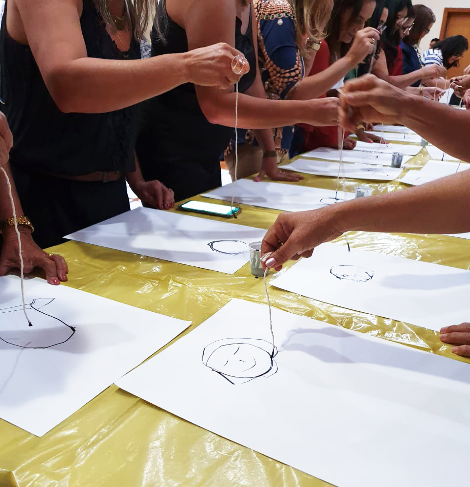 Cerca de 10 pessoas seguram pedaços de barbante e contornam ilustrações feitas em folhas de papel, que estão apoiadas em uma mesa retangular. Fim da descrição.