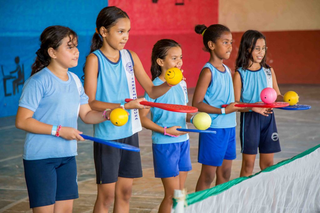 Em uma quadra, cinco meninas trajando uniformes seguram pequenas raquetes de tênis coloridas e bolas amarelas. Fim da descrição.