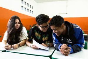 Em sala de aula, três estudantes sentados sorriem, observando uma página do caderno. Fim da descrição.