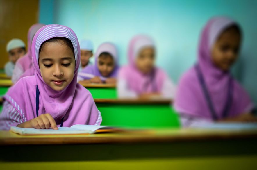 Em primeiro plano, dentro da sala de aula, uma menina usando hijab (conjunto de vestimentas de origem islâmica) faz lição, apoiando seu caderno em uma carteira escolar. Ao fundo, outras cinco crianças realizam a mesma ação. Fim da descrição.