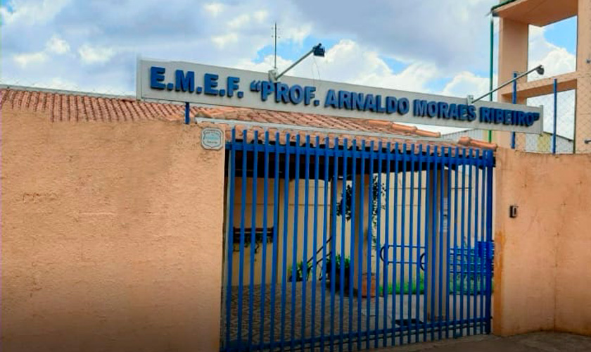 Fachada da escola Prof. Arnaldo Moraes Ribeiro, que tem muros amarelos e portão azul. Fim da descrição.