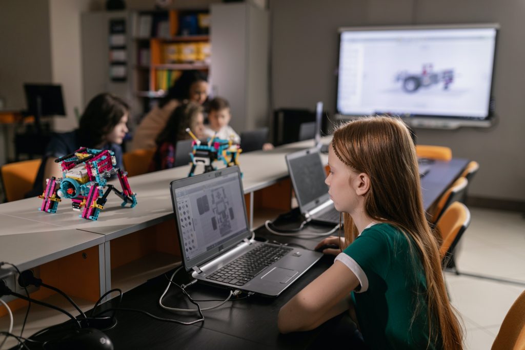 Estudantes em ambiente escolar com vários computadores. Em primeiro plano, uma estudante utiliza um computador de mesa. Ao fundo, outros três estudantes interagem entre si. Nas mesas há também protótipos robóticos. Fim da descrição.