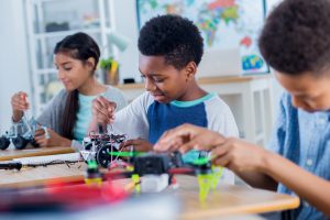 Fotografia de três estudantes, uma menina e dois meninos, em laboratório escolar, que manuseiam equipamentos de robótica e protótipos eletrônicos compostos por fios, ferragens e rodas; estes objetos estão apoiados sobre uma mesa. Fim da descrição.