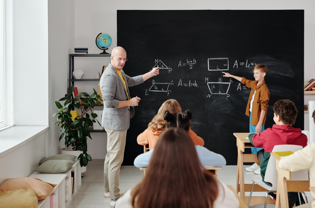 Cinco estudantes em sala de aula. Eles aparecem sentados, com exceção de um menino que está em pé, ao lado de um adulto. Os dois apontam para uma lousa, na qual estão desenhadas figuras geométricas e expressões matemáticas. Fim da descrição.