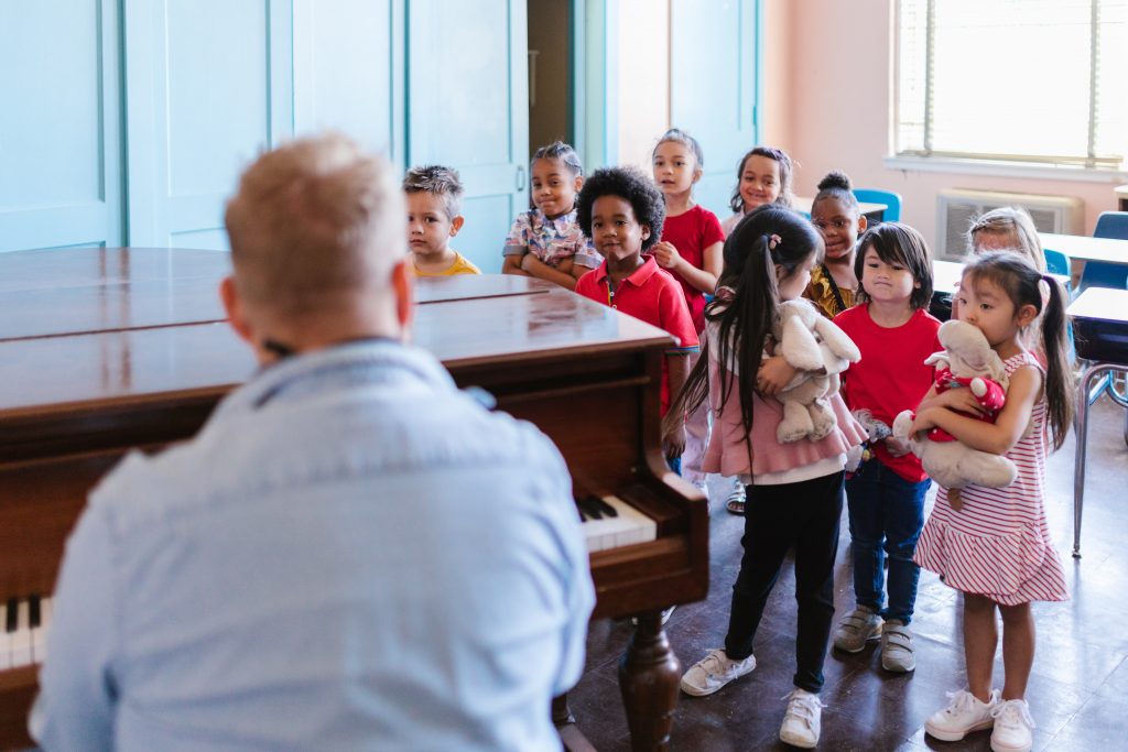 Em ambiente escolar, adulto toca piano olhando para um grupo de crianças. Elas também o observam. Fim da descrição.