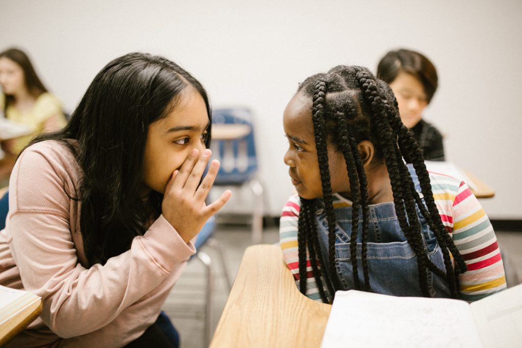 Duas estudantes, uma parda e uma negra, estão em destaque em uma sala de aula. Uma delas está conversando com a mão escondendo a boca, enquanto a outra a olha atentamente. Fim da descrição.