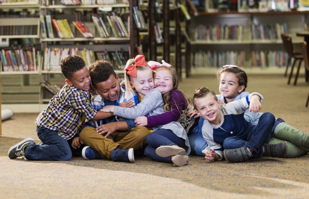 Grupo de seis crianças sorridentes, sendo três meninas brancas e dois meninos negros e um branco, sentadas se abraçando em uma biblioteca escolar. Fim da descrição. 