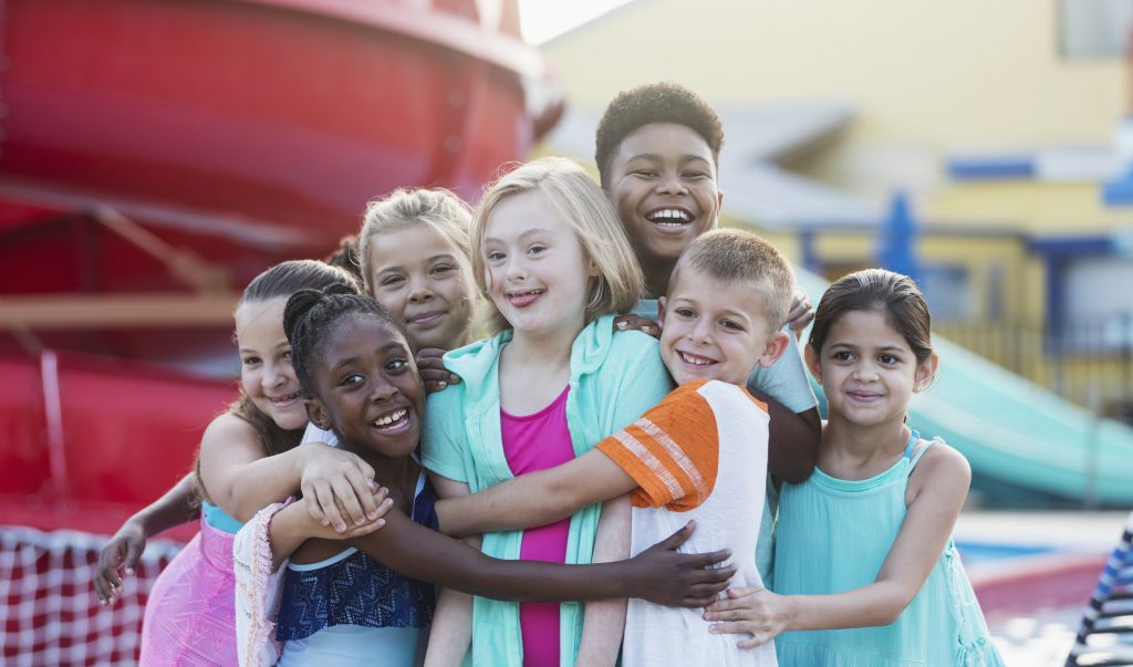 Foto com um grupo de sete crianças, meninas, sendo três brancas, duas pardas e uma negra, e dois meninos, um branco e um negro, sorrindo e se abraçando em um espaço aberto. A menina do meio tem síndrome de down.