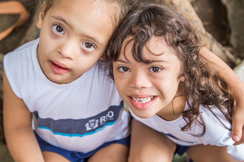 Na fotografia, um menino branco e uma menina branca, ambos com Síndrome de Down, estão se abraçando e sorrindo olhando para frente. Fim da descrição. 