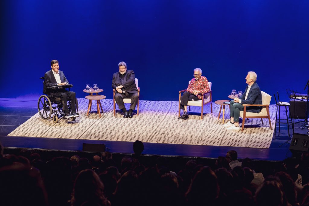Fotografia de três homens brancos e um homem negro sobre um palco. Todos estão sentados, um deles em cadeira de rodas. O palco tem cenário de fundo azul escuro. Fim da descrição. 
