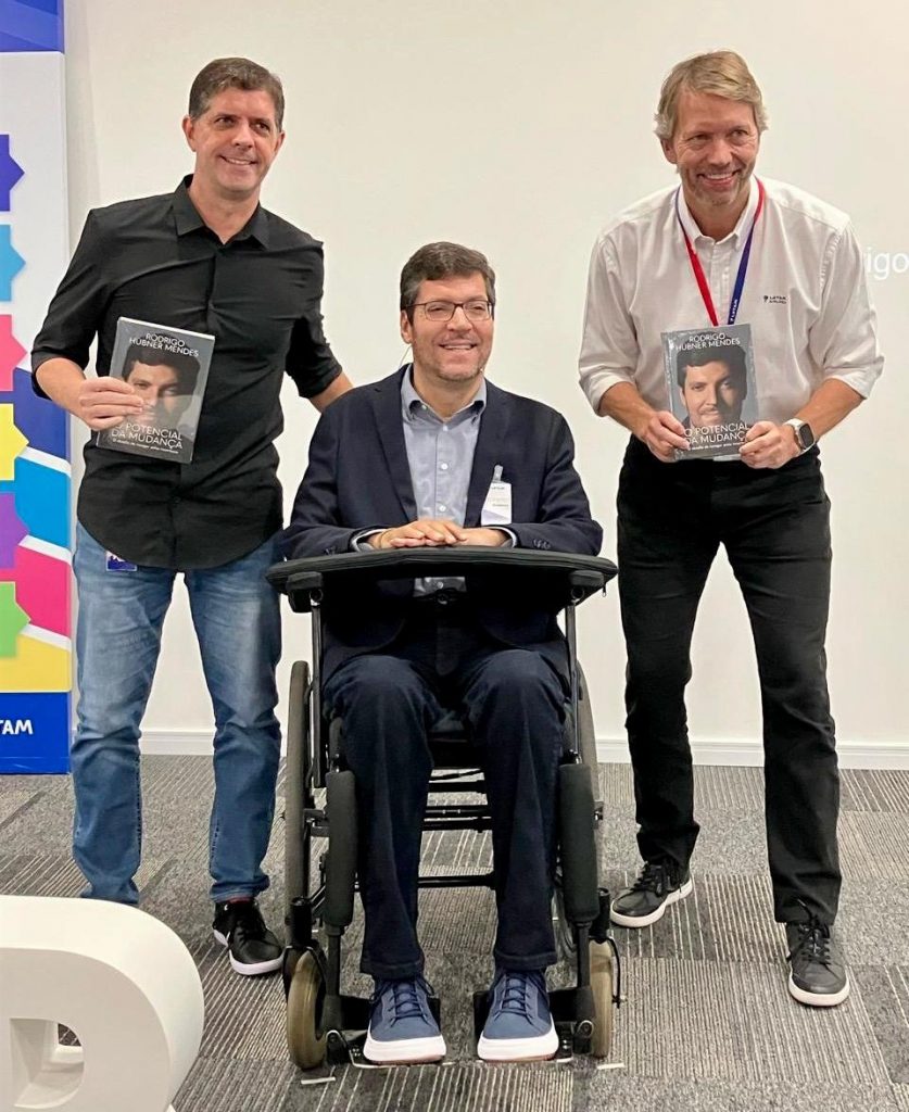 Três homens brancos, sendo um em cadeira de rodas e os outros dois em pé ao seu lado, ambos seguram um exemplar do livro "O Potencial da Mudança". Fim da descrição.  