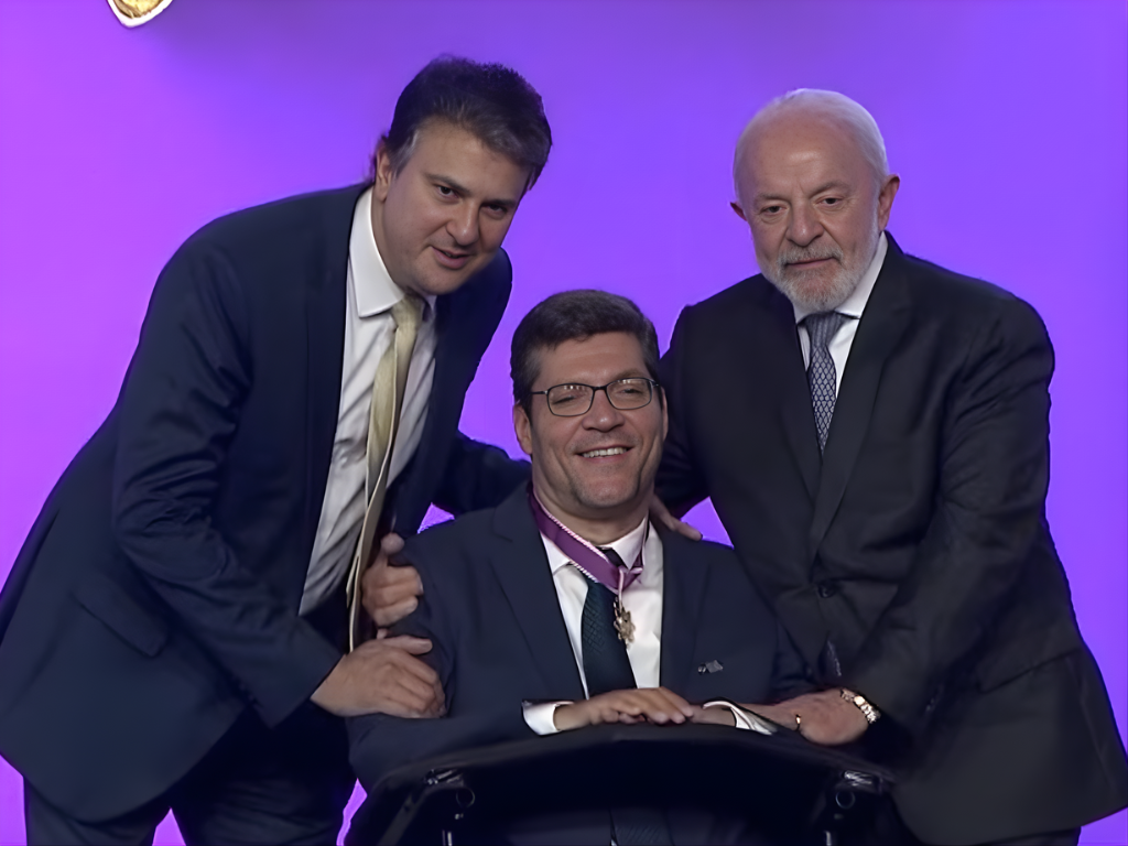 Foto do superintendente do Instituto Rodrigo Mendes, Rodrigo Hübner Mendes, que é cadeirante, ao lado do presidente da República Luís Inácio Lula da Silva, e do ministro da Educação Camilo Santana. Os três são homens brancos, usam terno escuro e gravata, e estão sorrindo para a foto em um fundo lilás. Fim da descrição.