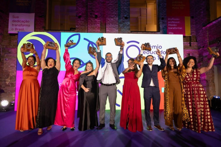 Grupo de nove pessoas em um palco, posando lado a lado e sorrindo, todas com troféus erguidos acima da cabeça. Estão vestidas com roupas formais e coloridas em um ambiente iluminado com luzes roxas e quentes. Ao fundo, há um painel grande com a identidade visual do Prêmio Educado Nota 10 e banners com mensagens sobre educação, indicando uma cerimônia de premiação.